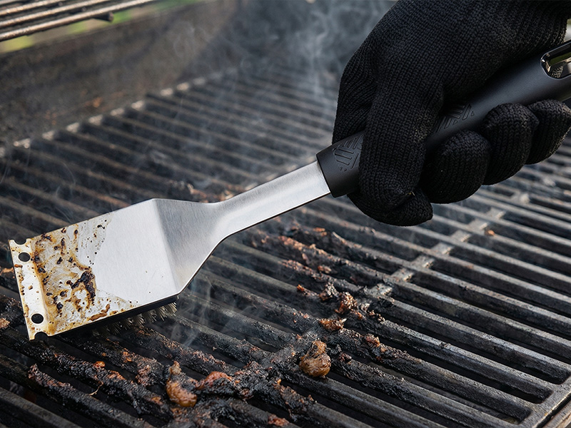 L-TOOLS hand cleaning a grill with a scraper, emphasizing maintenance and hygiene of cooking equipment.