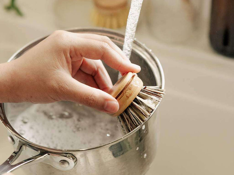 L-TOOLS person using a wooden brush to clean a pot, emphasizing natural and effective cleaning tools.